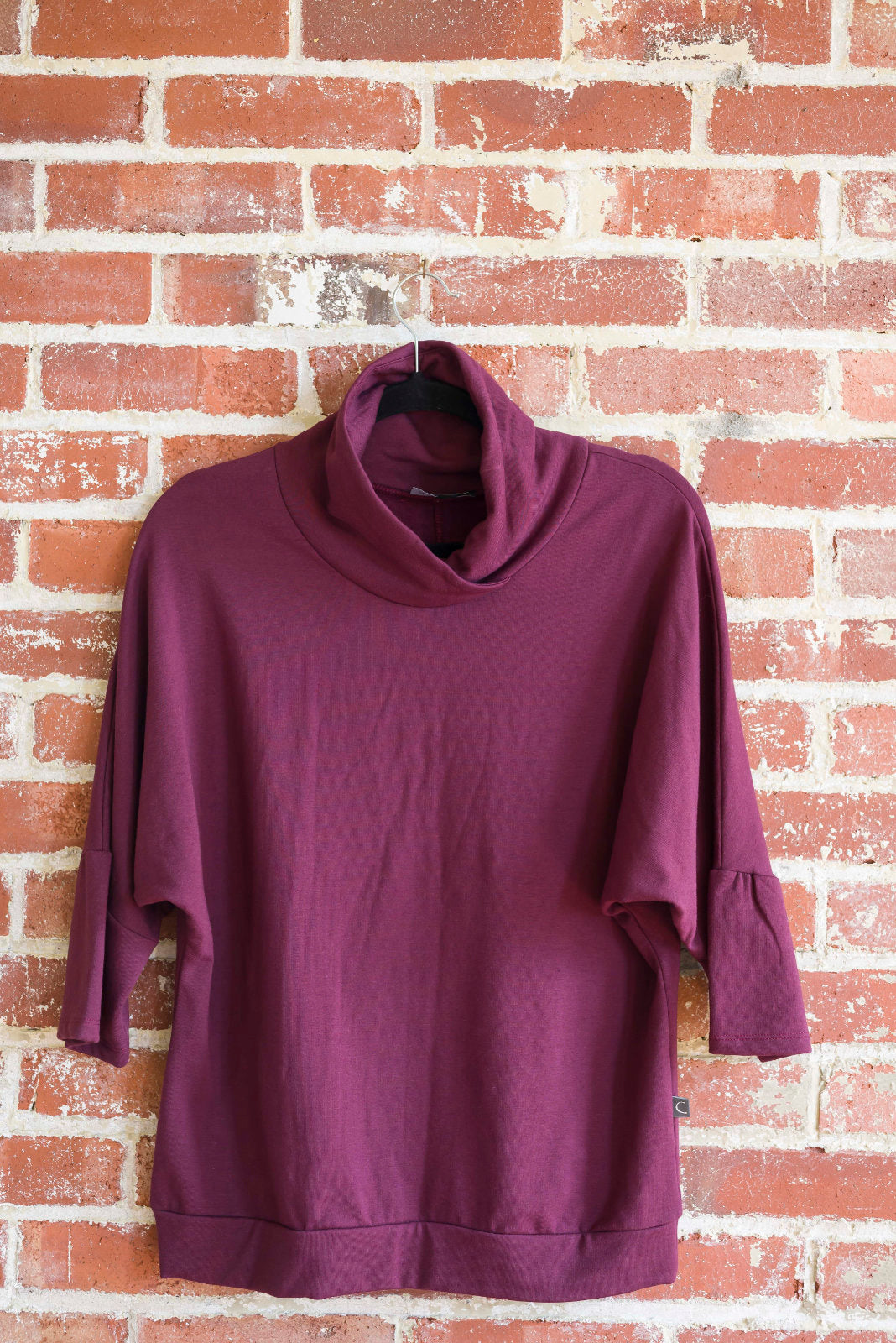 The BeAAA sweater by Marie C in Wine on a hanger against a brick wall
