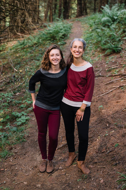 Two women wearing the Jaja Top by Marie C, one in Black and one in Wine. It's a sweatshirt with contrasting fabric bands at the wide neck, 7/8 sleeves, and hem. They are standing on a forest path.