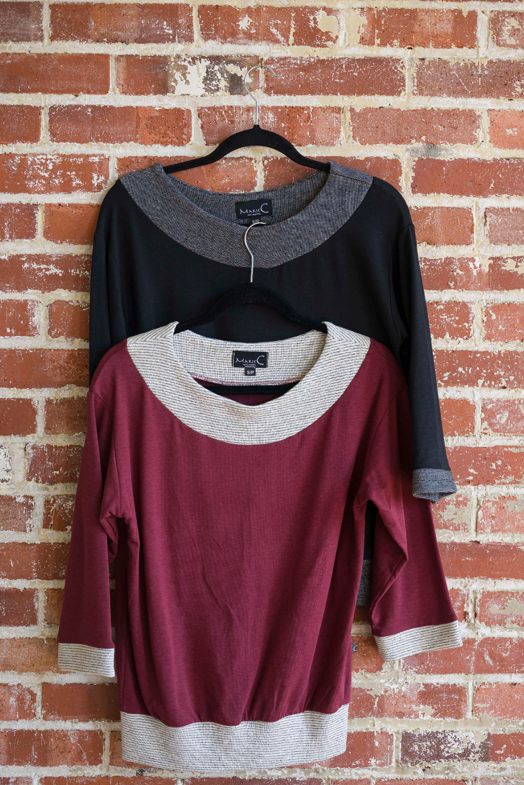 The Jaja Top by Marie C, one in Black and one in Wine, are shown hanging in front of a brick wall. It's a sweatshirt with contrasting fabric bands at the wide neck, 7/8 sleeves, and hem. 