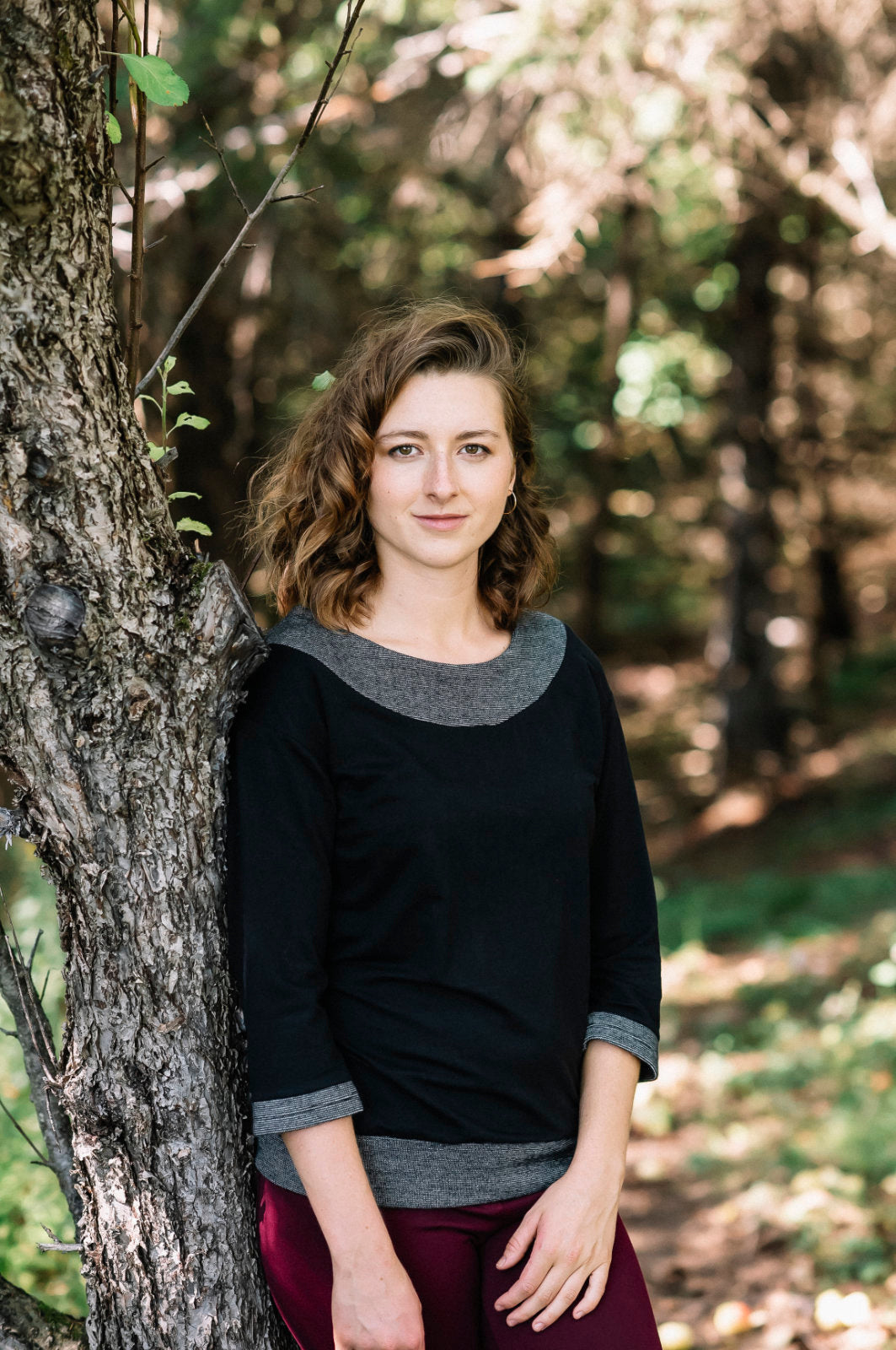 A woman wearing the Jaja Top by Marie C in Black. It's a sweatshirt with contrasting fabric bands at the wide neck, 7/8 sleeves, and hem. She is standing in front of a tree. 