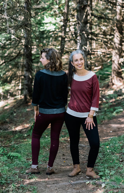 Two women wearing the Jaja Top by Marie C, one in Black and one in Wine. It's a sweatshirt with contrasting fabric bands at the wide neck, 7/8 sleeves, and hem. They are standing on a forest path.