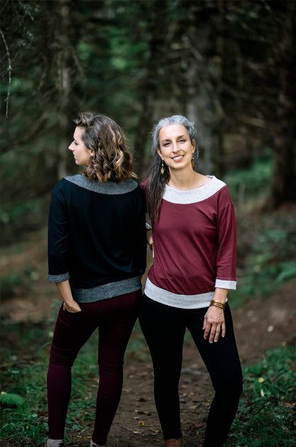Two women wearing the Jaja Top by Marie C, one in Black and one in Wine. It's a sweatshirt with contrasting fabric bands at the wide neck, 7/8 sleeves, and hem. They are standing on a forest path.