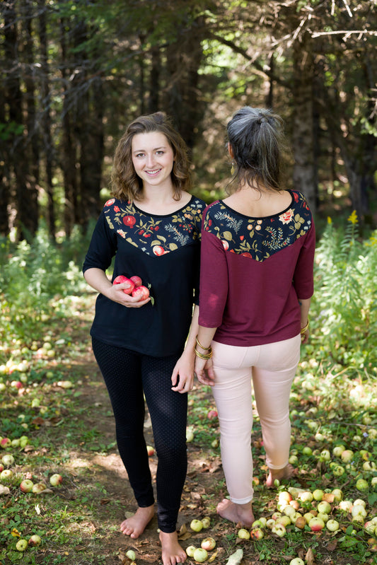 Two women wearing the Manzana Top by Marie C, one in Black and one in Wine. It's a reversible top with autumn print fabric along the neck on one side, and 3/4 sleeves. They are standing in in an apple orchard. 