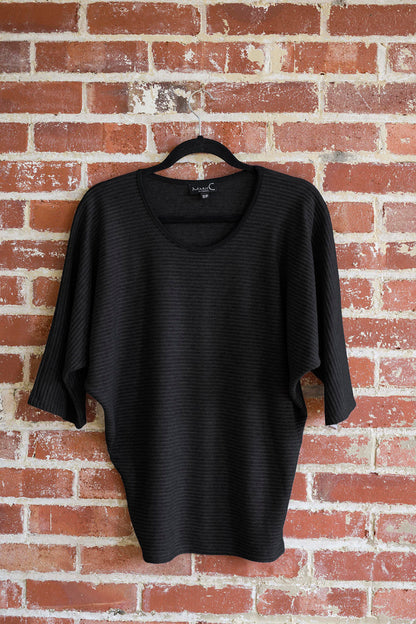 The Perla Top by Marie C in Black, with a wide round neck, 3/4 batwing sleeves, and fitted waist in ribbed knit fabric, is shown hanging against a brick wall. 