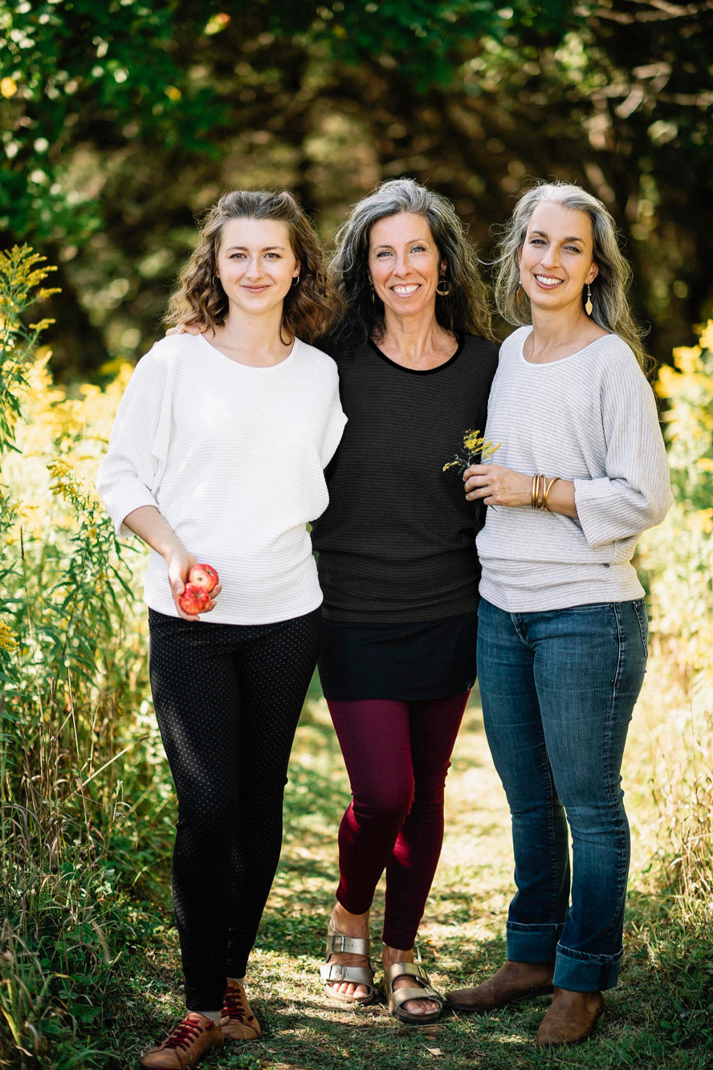Three women wearing the Perla Top by Marie C,  one in Ivory and one in Black and one in stripes, with a wide round neck, 3/4 batwing sleeves, and fitted waist in ribbed knit fabric. They are standing outside. 