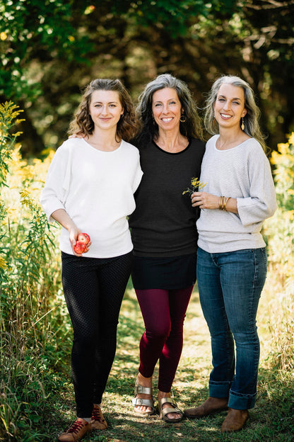 Three women wearing the Perla Top by Marie C,  one in Ivory and one in Black and one in stripes, with a wide round neck, 3/4 batwing sleeves, and fitted waist in ribbed knit fabric. They are standing outside. 