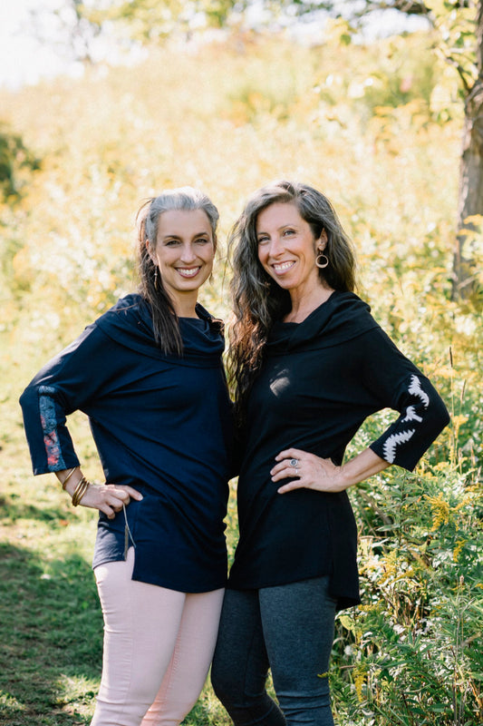 Two women wearing the Tunic Java by Marie C, one in Black and one in Navy, featuring a cowl collar, 7/8 batwing sleeves with contrast fabric, and a hi-low hemline with a slit detail. They are standing in a forest. 