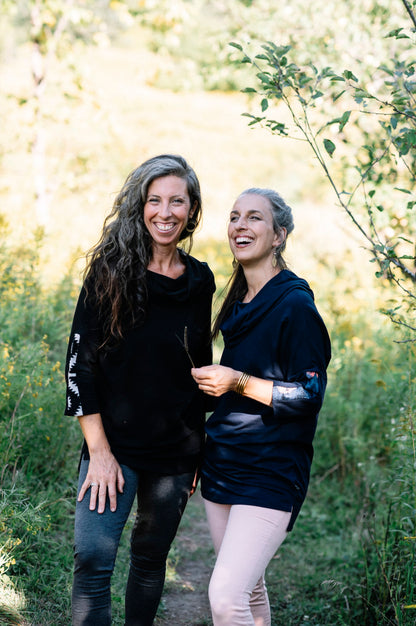 Two women wearing the Tunic Java by Marie C, one in Black and one in Navy, featuring a cowl collar, 7/8 batwing sleeves with contrast fabric, and a hi-low hemline with a slit detail. They are standing in a forest. 