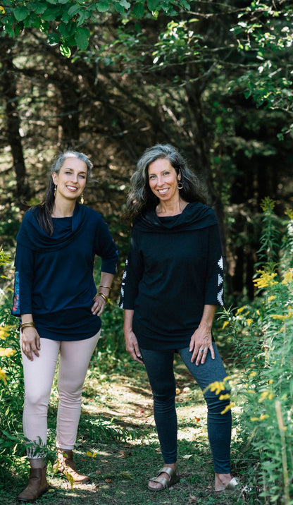 Two women wearing the Tunic Java by Marie C, one in Black and one in Navy, featuring a cowl collar, 7/8 batwing sleeves with contrast fabric, and a hi-low hemline with a slit detail. They are standing in a forest. 