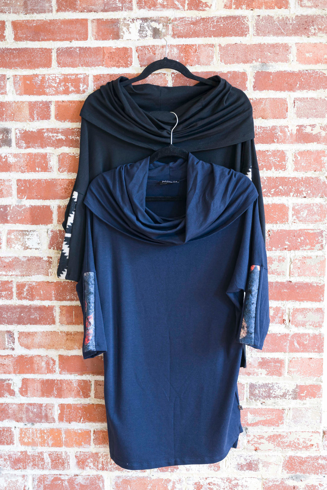 The unic Java by Marie C, one in Black and one in Navy, are shown hanging against a brick wall. It features a cowl collar, 7/8 batwing sleeves with contrast fabric, and a hi-low hemline with a slit detail. 