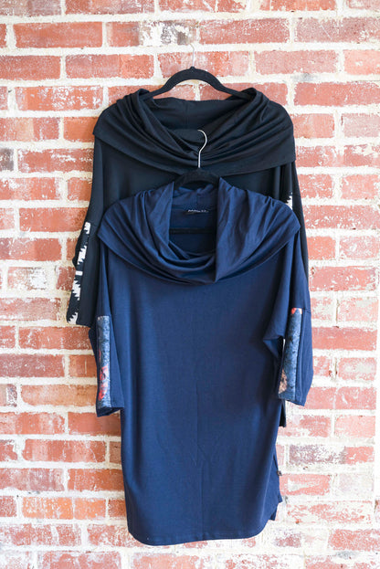 The unic Java by Marie C, one in Black and one in Navy, are shown hanging against a brick wall. It features a cowl collar, 7/8 batwing sleeves with contrast fabric, and a hi-low hemline with a slit detail. 