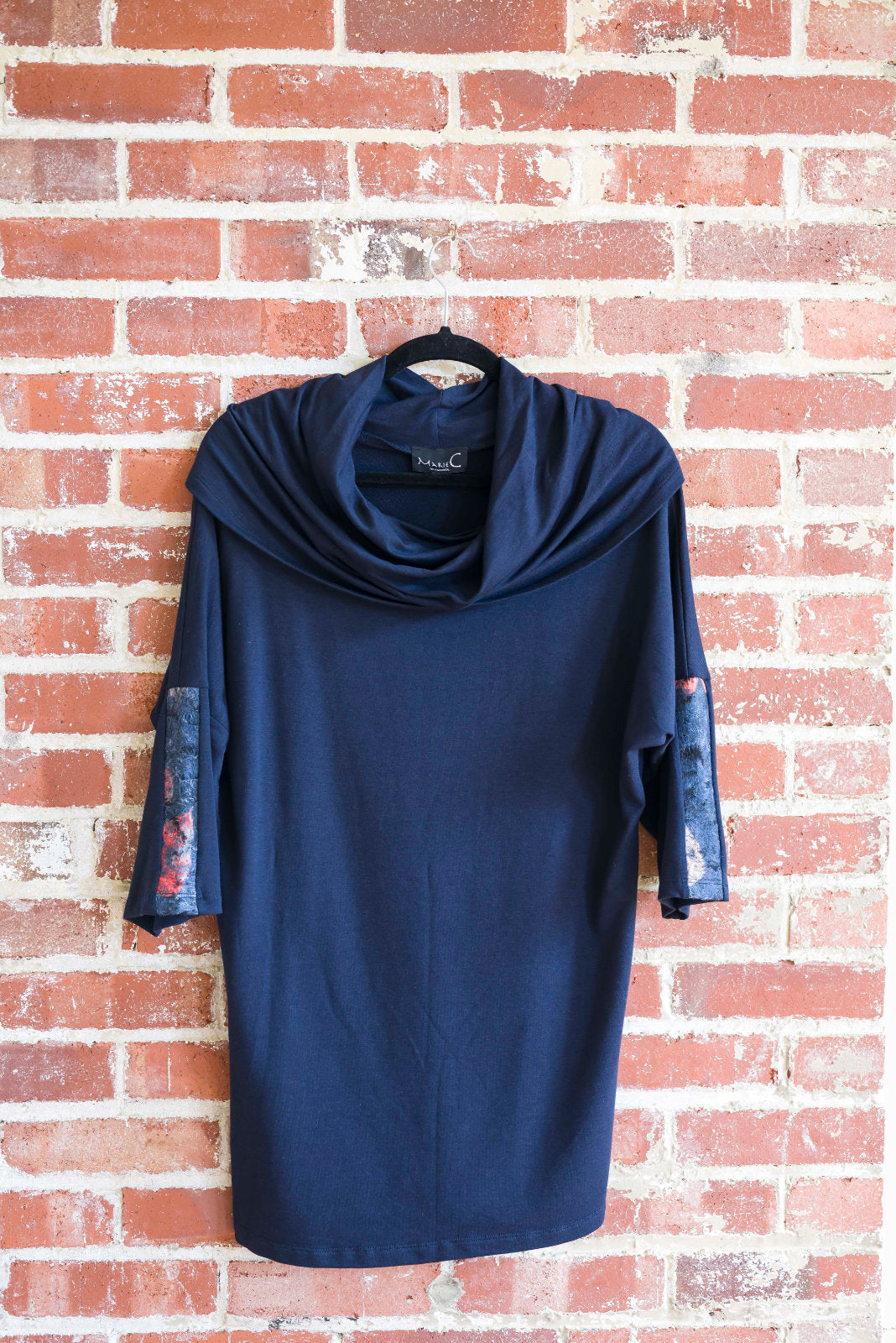 The tunic Java by Marie C in Navy is shown hanging against a brick wall. It features a cowl collar, 7/8 batwing sleeves with contrast fabric, and a hi-low hemline with a slit detail. 