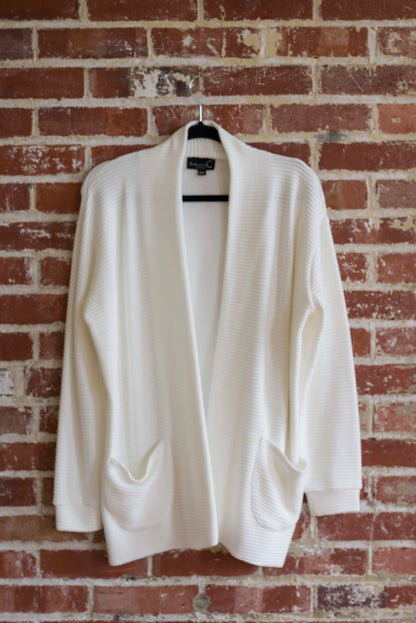 The Margo Cardigan by Marie C in Ivory, a long open cardigan  in ribbed knit with rounded patch pockets and dropped sleeves, is shown hanging in front of a brick wall. 