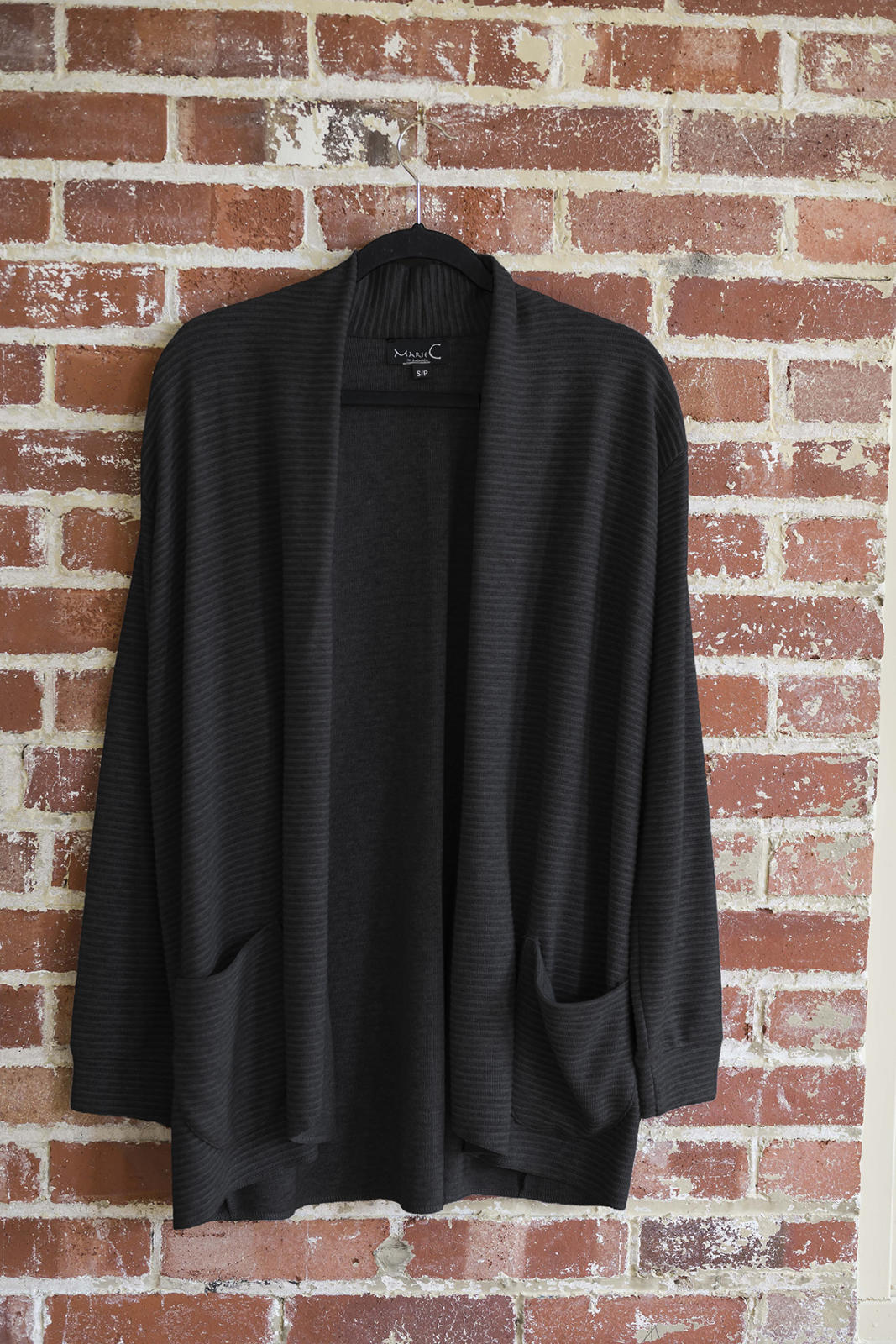 The Margo Cardigan by Marie C in Black, a long open cardigan  in ribbed knit with rounded patch pockets and dropped sleeves, is shown hanging in front of a brick wall. 