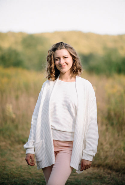 A woman wearing the Perla Top by Marie C in Ivory, with a wide round neck, 3/4 batwing sleeves, and fitted waist in ribbed knit fabric. She is wearing it with the matching Margo Cardigan and pink jeans and is standing outside. 