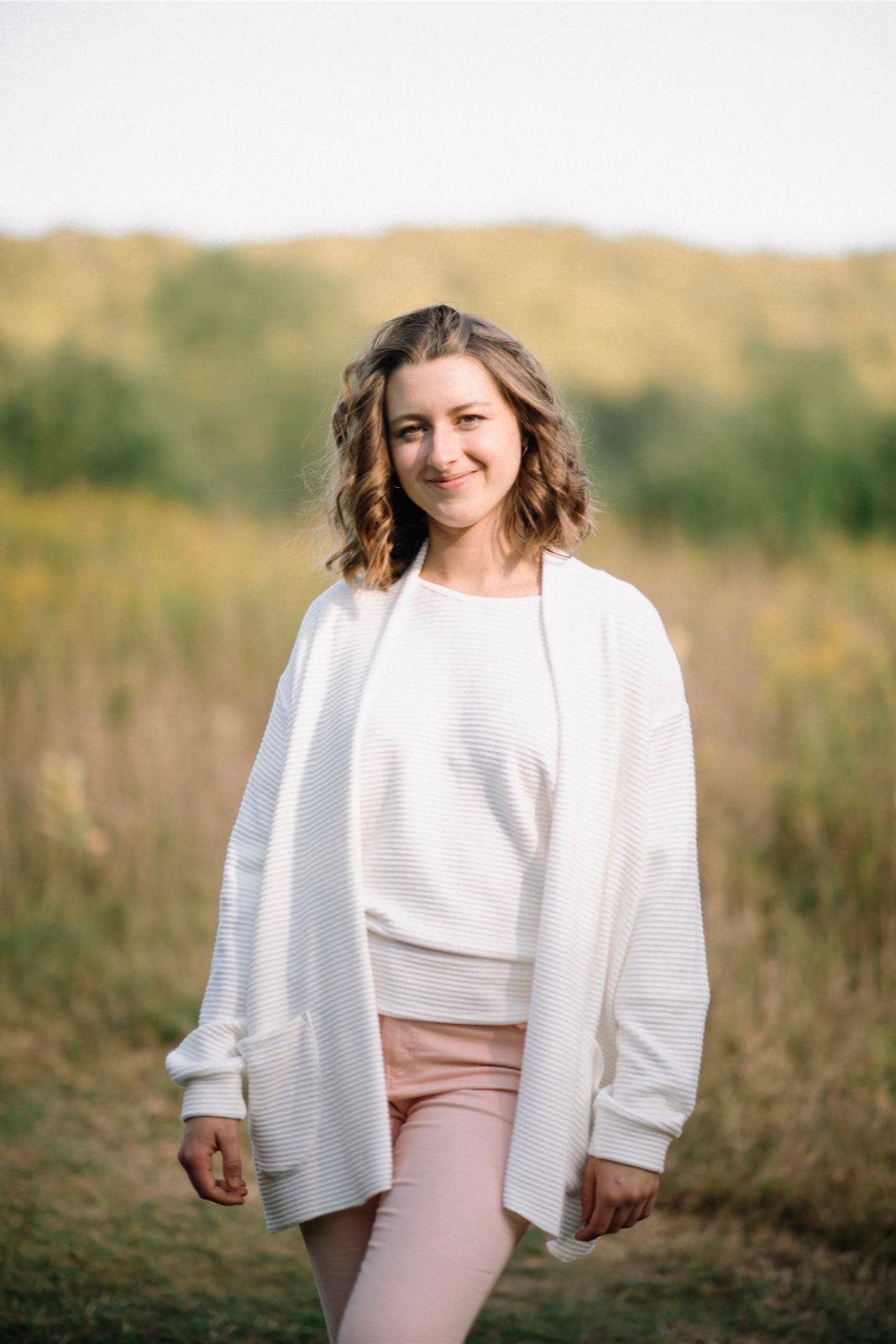 A woman wearing the Margo Cardigan by Marie C in Ivory, a long open cardigan  in ribbed knit with rounded patch pockets and dropped sleeves. She is wearing it with pink jeans and standing in a field. 