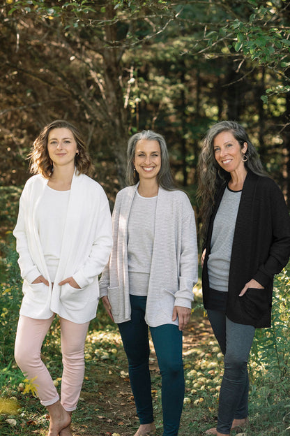 Three women wearing the Margo Cardigan by Marie C in Ivory, stripes, and black, a long open cardigan  in ribbed knit with rounded patch pockets and dropped sleeves. They are wearing them with jeans and standing in a forest.
