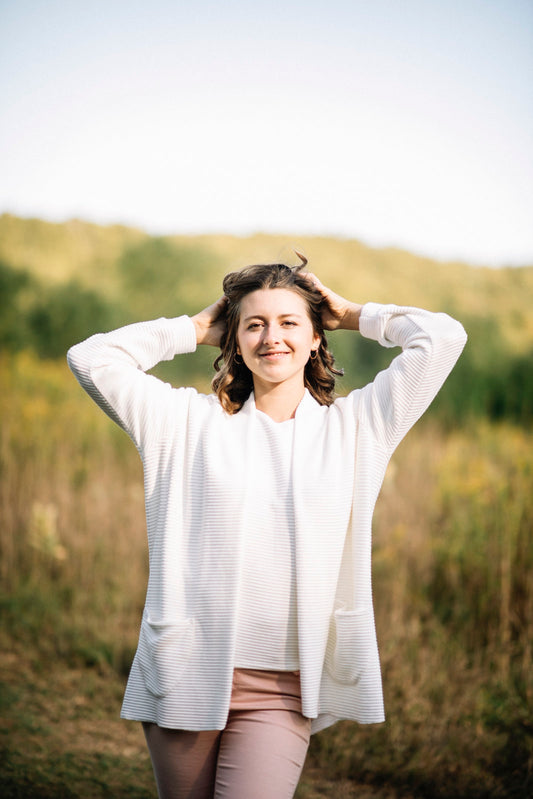 A woman wearing the Margo Cardigan by Marie C in Ivory, a long open cardigan  in ribbed knit with rounded patch pockets and dropped sleeves. She is wearing it with pink jeans and standing in a field. 