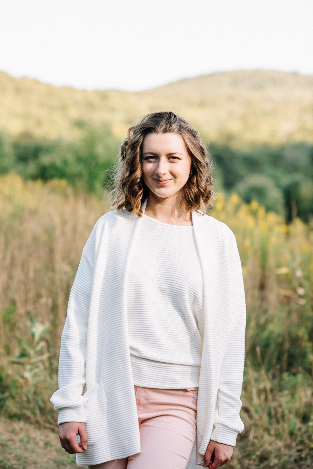 A woman wearing the Margo Cardigan by Marie C in Ivory, a long open cardigan  in ribbed knit with rounded patch pockets and dropped sleeves. She is wearing it with pink jeans and standing in a field. 