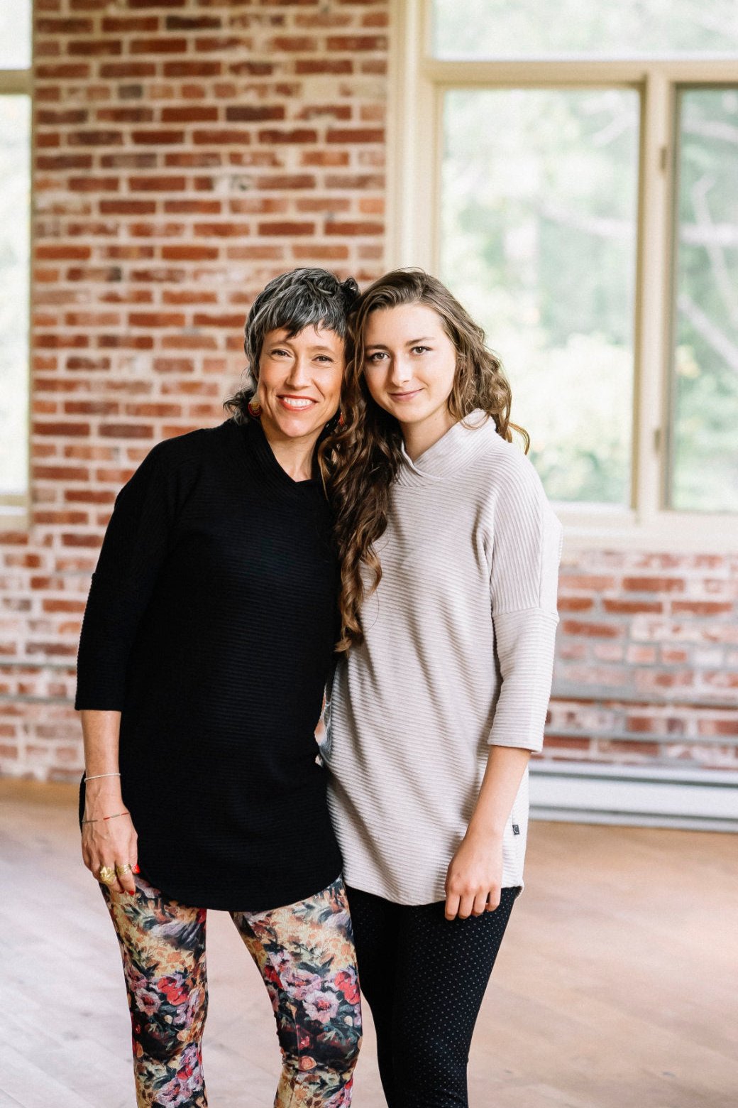 Two women wearing the Juliana Tunic by Marie C in Black and Sand, with a crossover neck, 3/4 sleeves, hi-low hemline, and ribbed fabric. They are standing in front of a brick wall. 