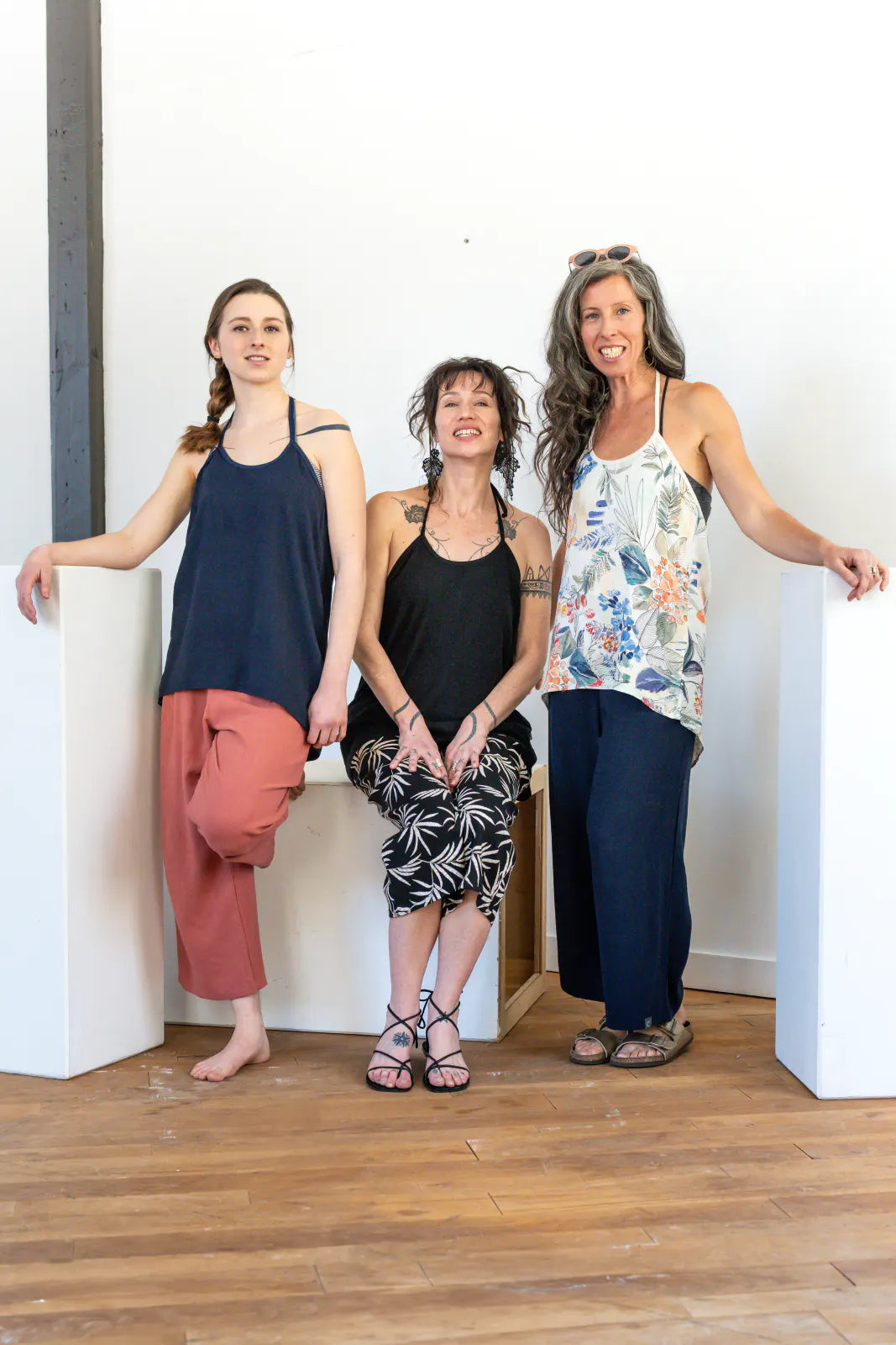 Three women wearing the Verano Pants by Marie C in various colours, they are wearing them with tank tops and standing in front of a white wall. 