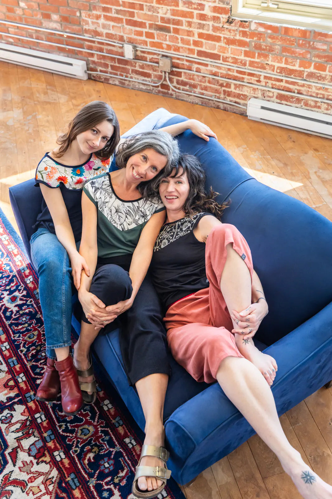 Three women wearing the pepita Top from Marie C, a reversible t-shirt with a printed fabric yoke on one side. From the left they are wearing it in 
Navy, Green, and Black  with Print.They are sitting on a blue couch in a room with brick walls and wooden floor.