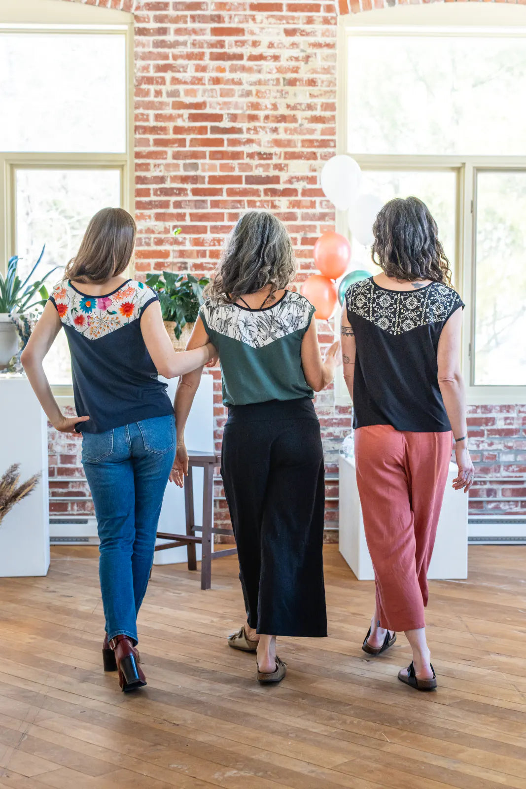 Back view of three women wearing the Pepita Top from Marie C, a reversible t-shirt with a printed fabric yoke on one side. From the left they are wearing it in 
Navy, Green, and Black with Print, and in a room with brick walls and balloons.