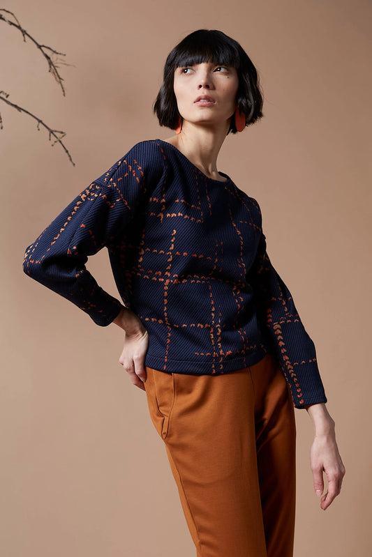 A woman wearing the Jackson Top by Melow in indigo with orange pants, standing in front of a peach background 