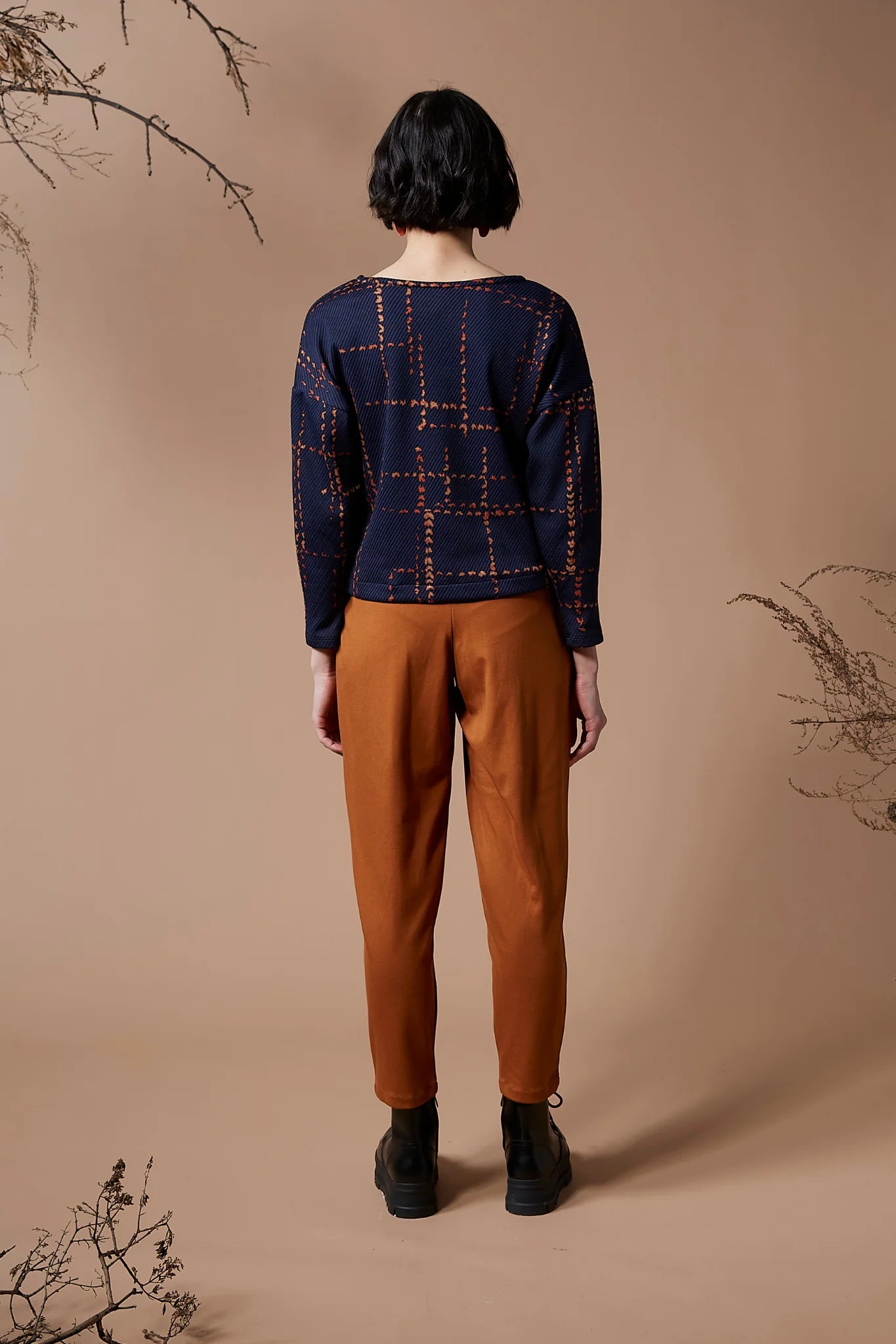 Back view of a woman wearing the Jackson Top by Melow in indigo with orange pants, standing in front of a peach background 