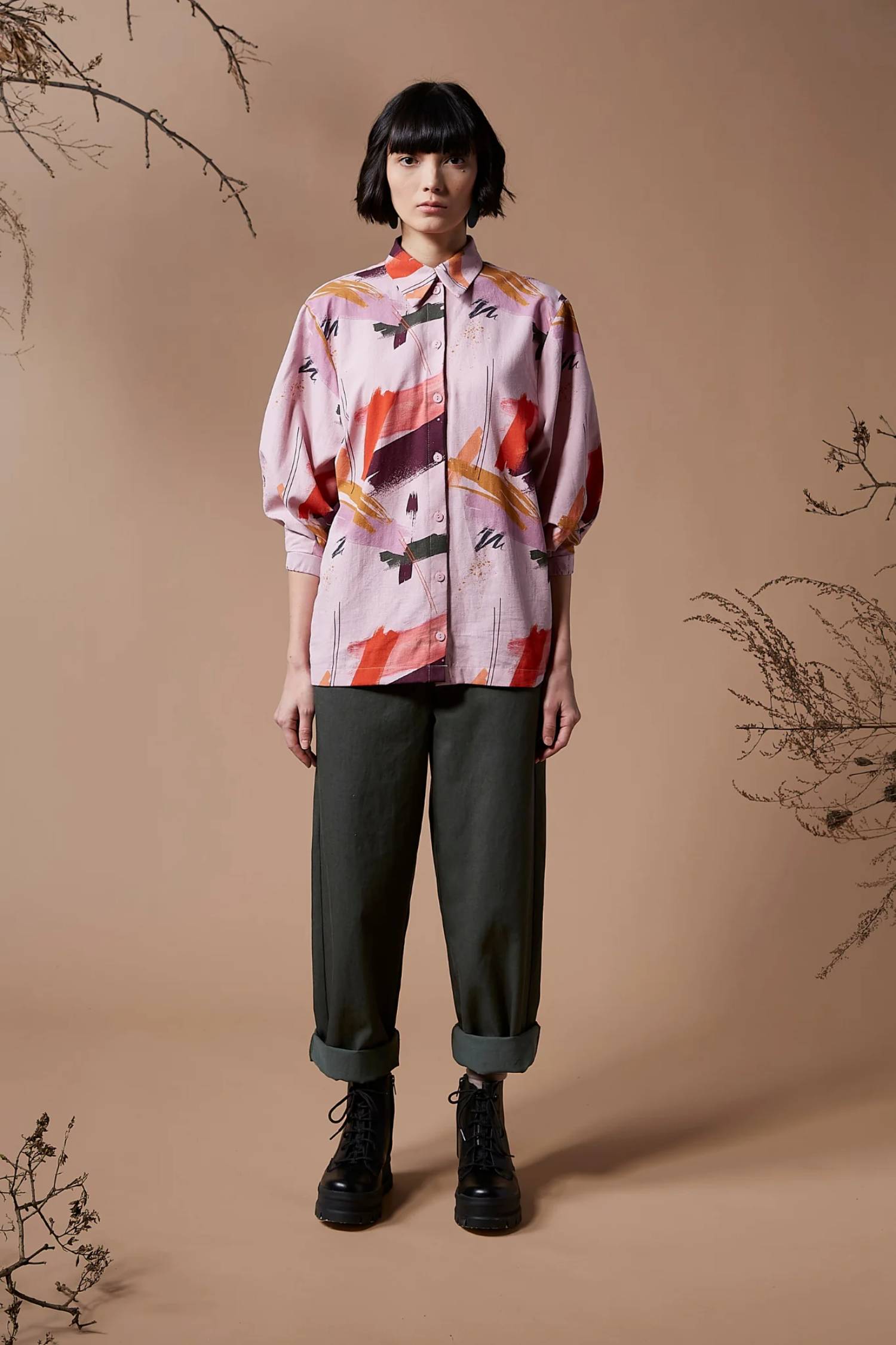 A woman wearing the Juliette Top by Melow in Sobel Print, featuring a pale pink background with splashes of colour.  The shirt has a blouse collar, button front, and 3/4 length puff sleeves. 