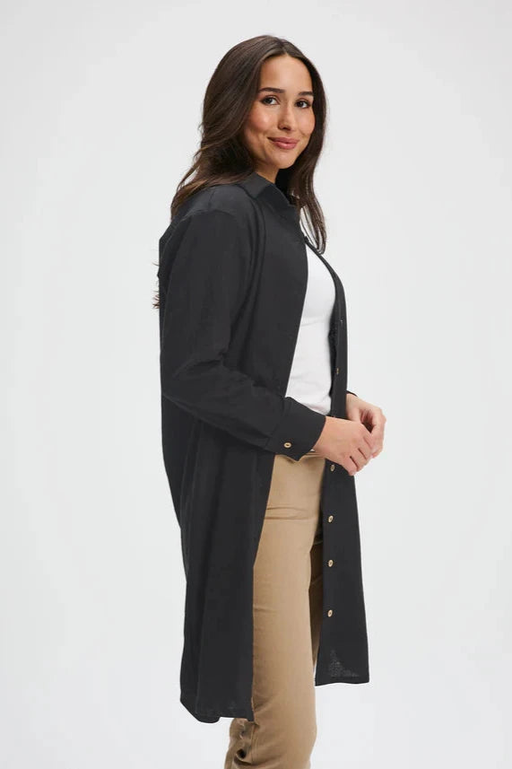 Side view of a woman wearing the Donnaconna Shirt Dress by Message Factory in Black, with a button front, classic collar, and rounded hem, made from natural ramie fabric. She is wearing it open over a white shirt and beige pants and is standing in front of a white background. 