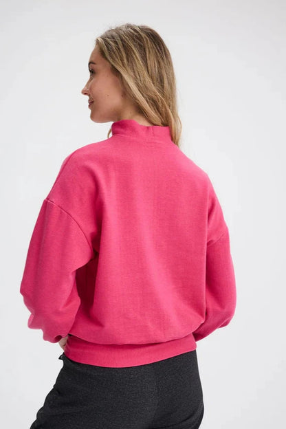 Back view of a woman wearing the Fermont Sweatshirt by Message Factory in Fuchsia, made from bamboo and cotton with a high neck, long sleeves with a puff detail at the cuffs, and fitted band at the bottom. She is wearing it with grey pants and is standing in front of a white background. 