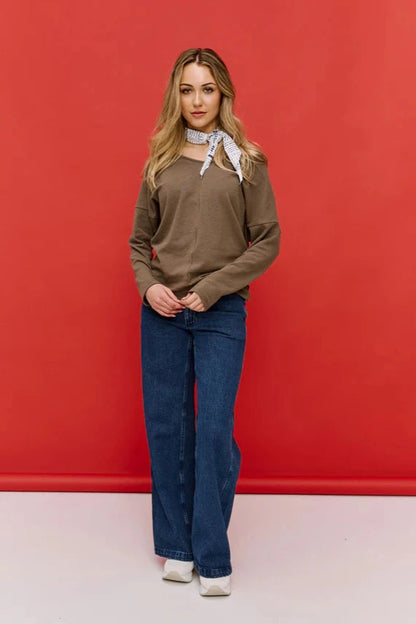 A woman wearing the Hochelaga Flared Leg Denim Pants by Message Factory, a wide leg jean made from organic cotton, with a brown sweater. She is standing in front of a red wall 