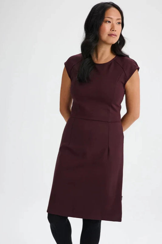 A woman wearing the Ivry Dress by Message Factory in Burgundy, a knee-length dress mad from a Lenzing Ecovero viscose blend, featuring a round neck and cap sleeves with princess seams and a defined waist. She is standing in front of a white background. 