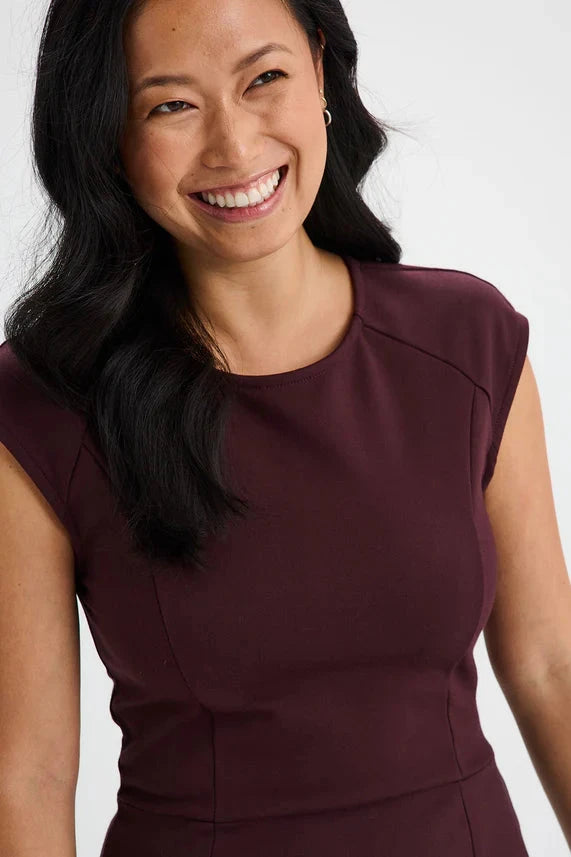 Close up of a woman wearing the Ivry Dress by Message Factory in Burgundy, a knee-length dress mad from a Lenzing Ecovero viscose blend, featuring a round neck and cap sleeves with princess seams and a defined waist. She is standing in front of a white background. 