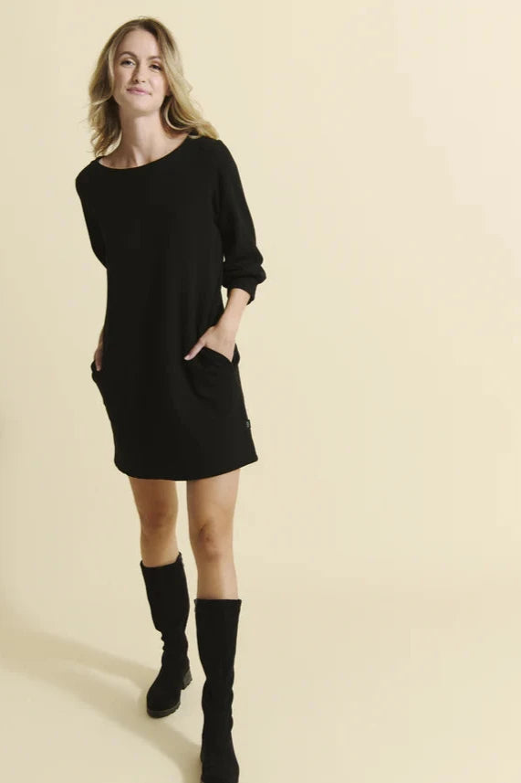 A woman wearing the Stukely Dress by Message Factory in Black, a semi-fitted bamboo/organic cotton fleece dress with a boat neck, 3/4 sleeves, side pockets, and a mid-thigh length. She is standing in front of a beige background. 