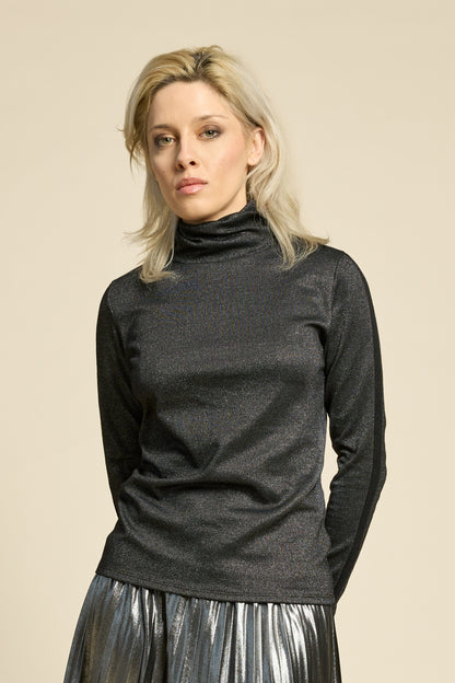 A woman wearing the Mortel Top by Cokluch in Facet Black, a metallic turtleneck with long sleeves. She is wearing it with the Feyer Skirt in Glow Knit Silver and standing in front of a beige background. 