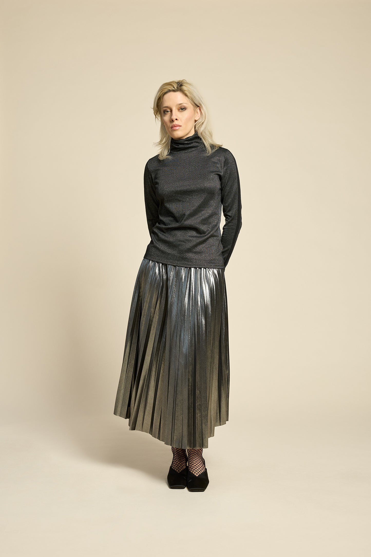 A woman wearing the Mortel Top by Cokluch in Facet Black, a metallic turtleneck with long sleeves. She is wearing it with the Feyer Skirt in Glow Knit Silver and standing in front of a beige background. 