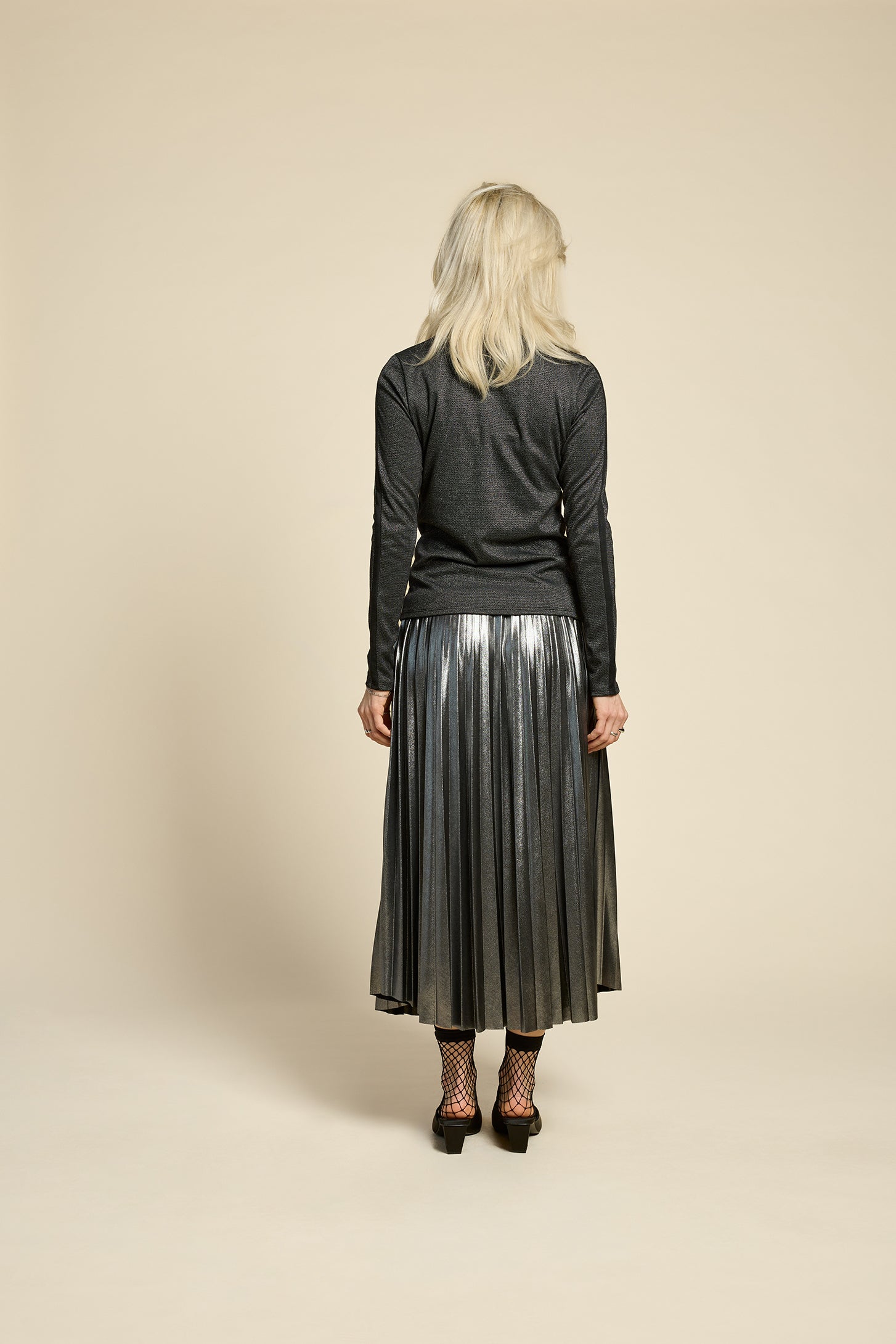 Back view of a woman wearing the Mortel Top by Cokluch in Facet Black, a metallic turtleneck with long sleeves. She is wearing it with the Feyer Skirt in Glow Knit Silver and standing in front of a beige background. 