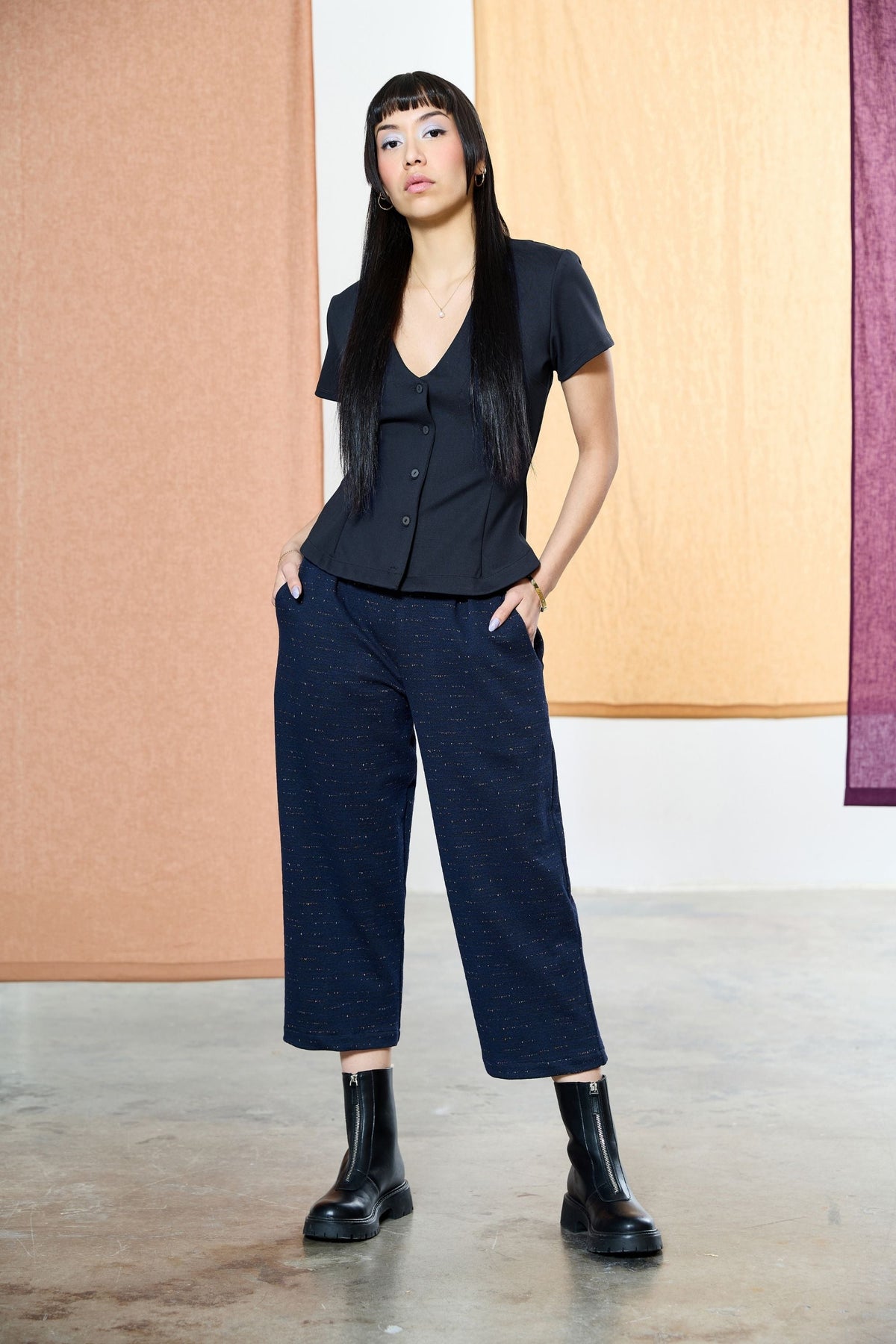 A woman wearing the Nathan Pants by Melow in Navy speckled fabric, 7/8 length wide legged pants with a high pull-on waist band and front pleats. She is wearing them with a black blouse and standing in front of a pastel background. 