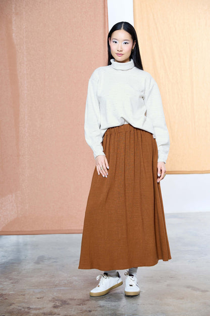 A woman wearing the Nathalie Top by Melow in Ivory, a relaxed fitting turtleneck with long barrel sleeves and a speckled texture. She is wearing it with a copper skirt and standing in front of a pastel background. 