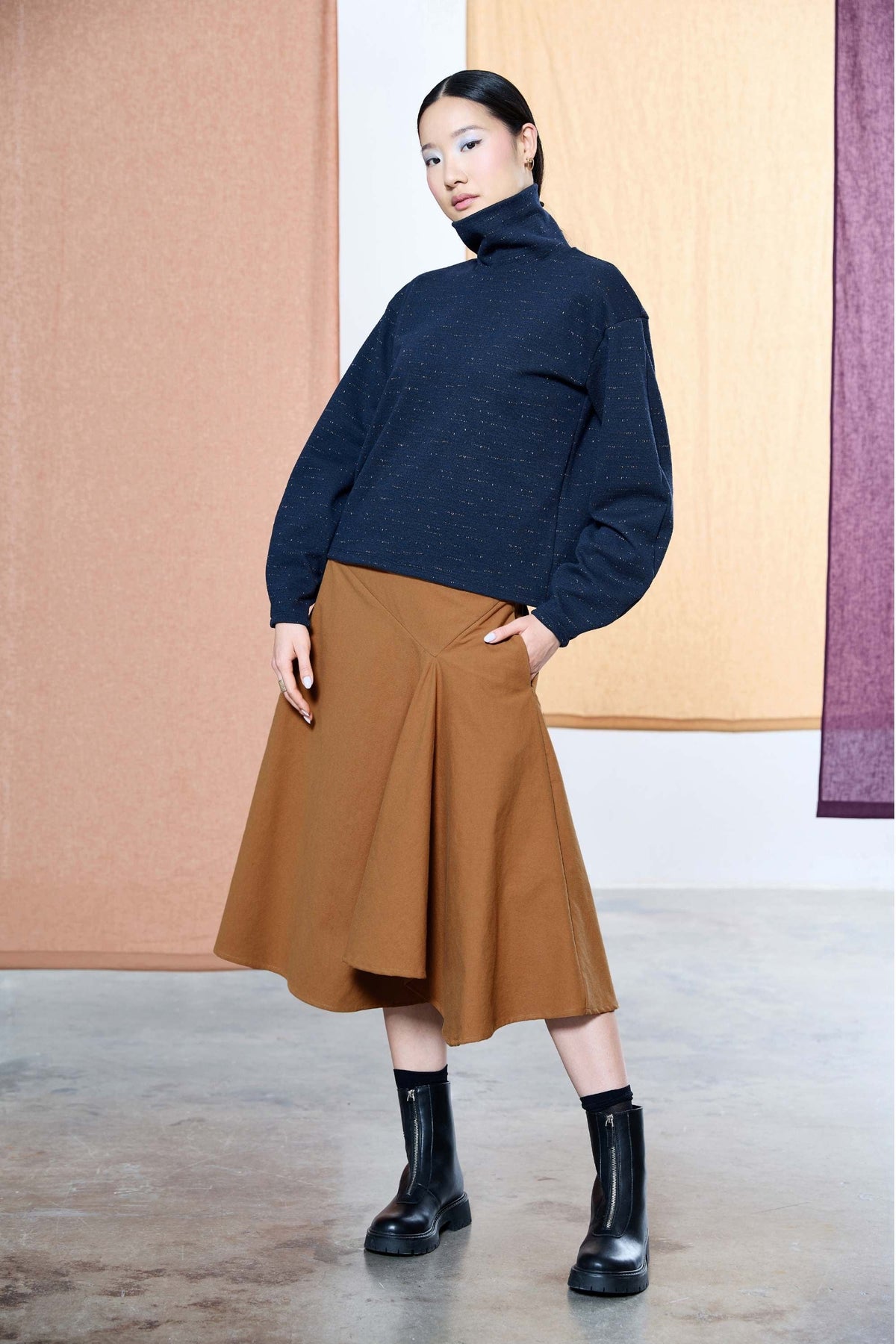 A woman wearing the Nathalie Top by Melow in Navy, a relaxed fitting turtleneck with long barrel sleeves and a speckled texture. She is wearing it with a copper skirt and standing in front of a pastel background. 