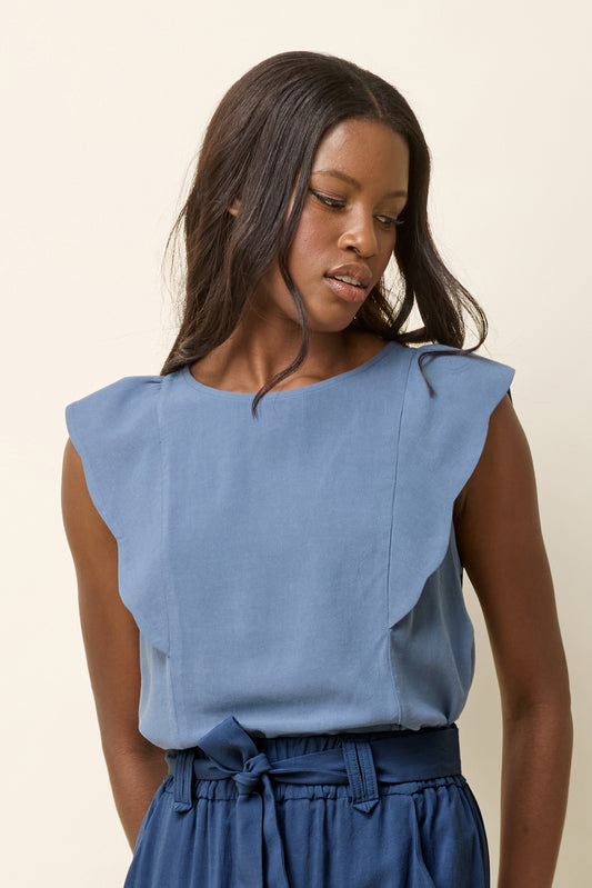 A woman wearing the Neema Top by Cokluch in Ocean, a light sleeveless top with ruffle details at the shoulders, a round neck, and a mid-hip length. She is wearing it with blue pants and standing in front of a beige background. 