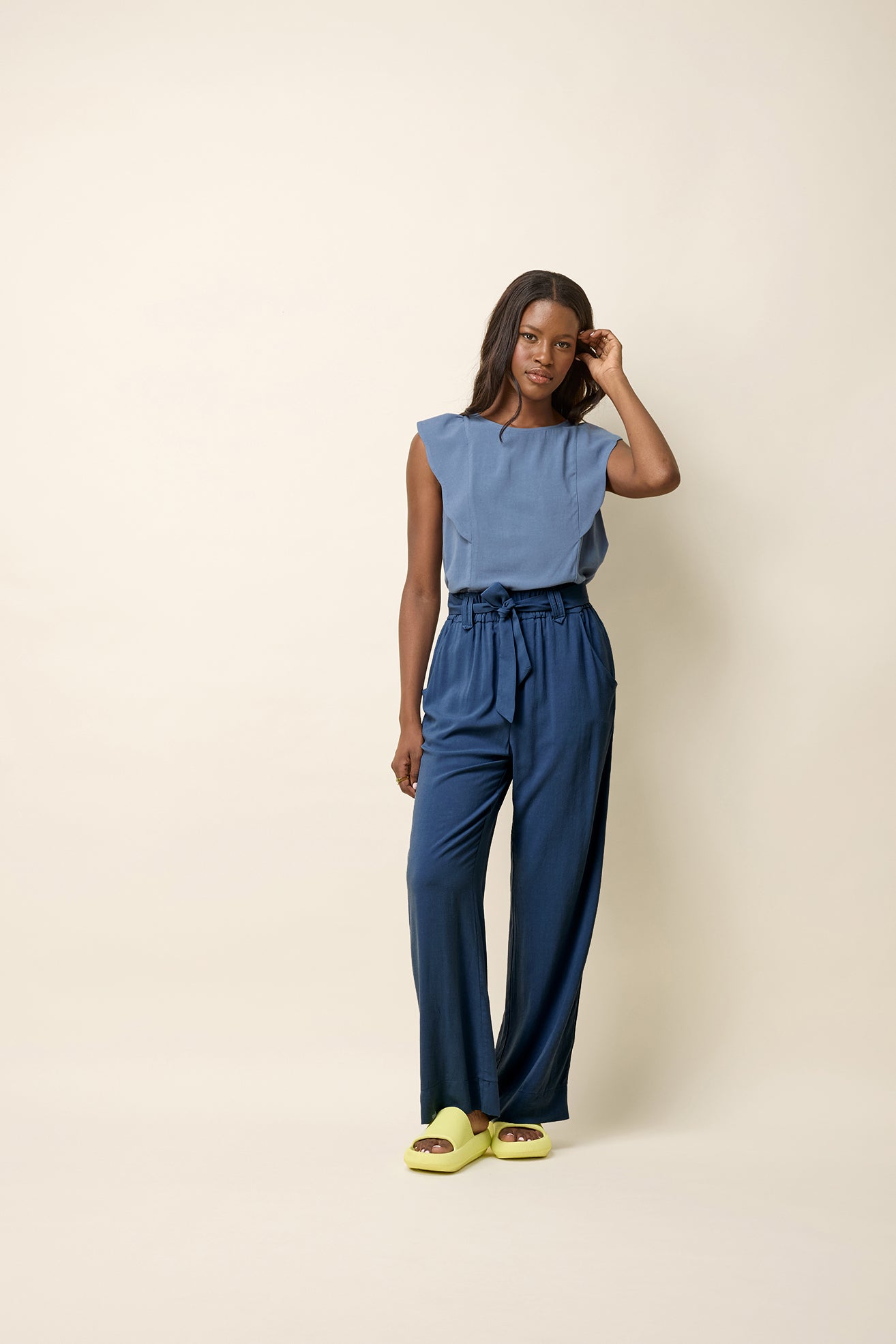 A woman wearing the Neema Top by Cokluch in Ocean, a light sleeveless top with ruffle details at the shoulders, a round neck, and a mid-hip length. She is wearing it with blue pants and standing in front of a beige background. 