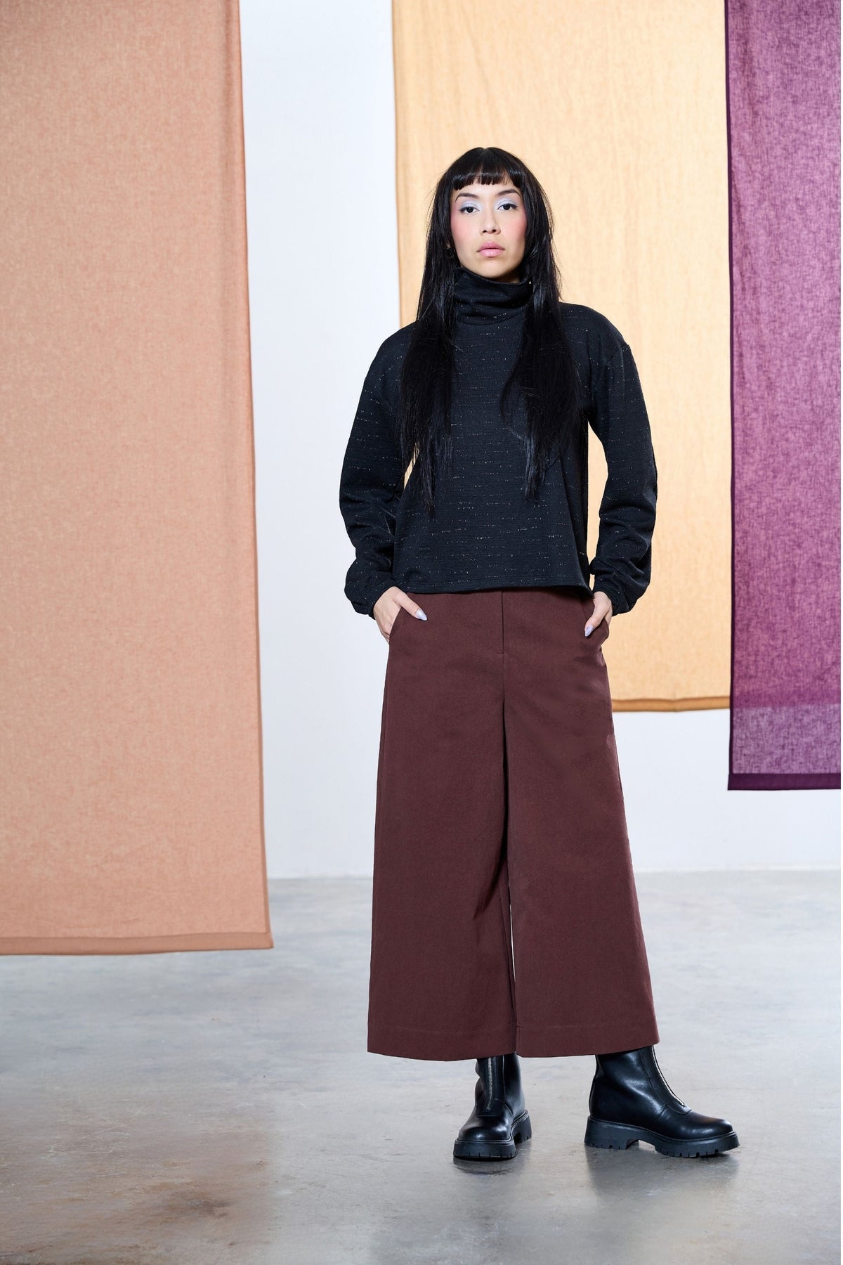  A woman wearing the Nigel Pants by Melow in Merlot, wide legged cropped pants with a high waist, elastic at the back, and button and zipper closure. She's wearing them with a black top and standing in front hanging fabric panels. 