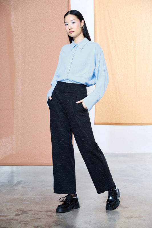 A woman wearing the Norbert Pants by Melow in Black speckled fabric, high waisted pull-one pants with a straight full length leg. She is wearing them with a light blue blouse and standing in front of a pastel background 