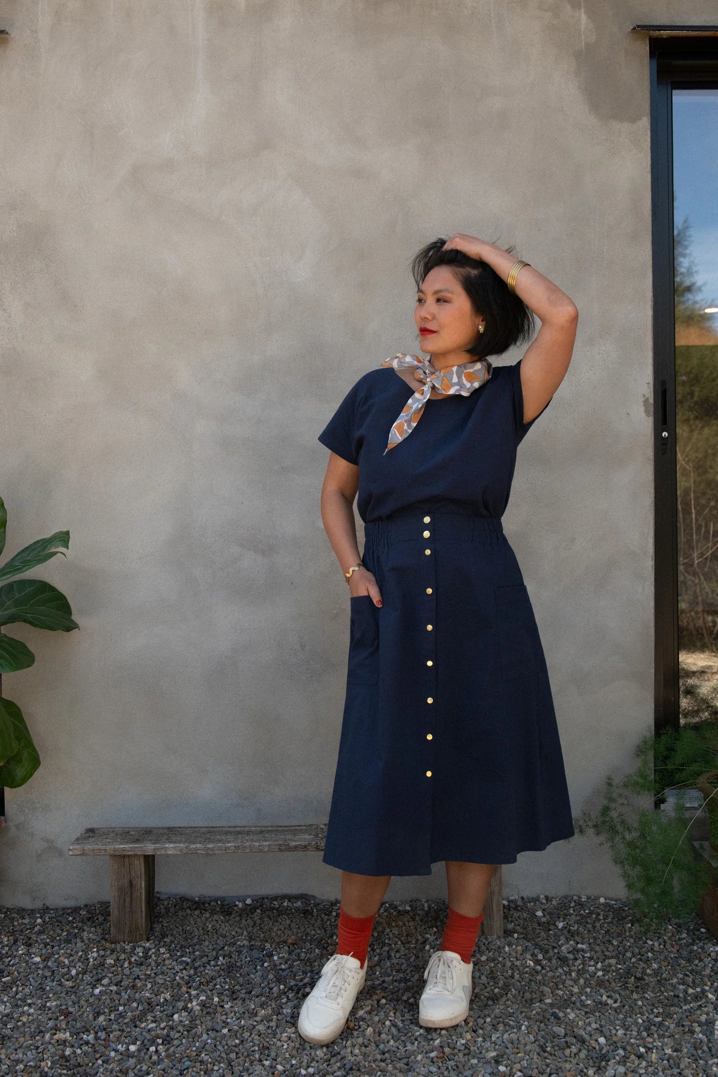 A woman wearing the Brendola Top from Kazak in Navy, a slightly cropped short sleeved top with three brass snaps that can be worn at the back or the front. She is wearing it with a scarf and the Nymphea skirt in Navy and standing outside in front of a grey wall with plants beside her. 
