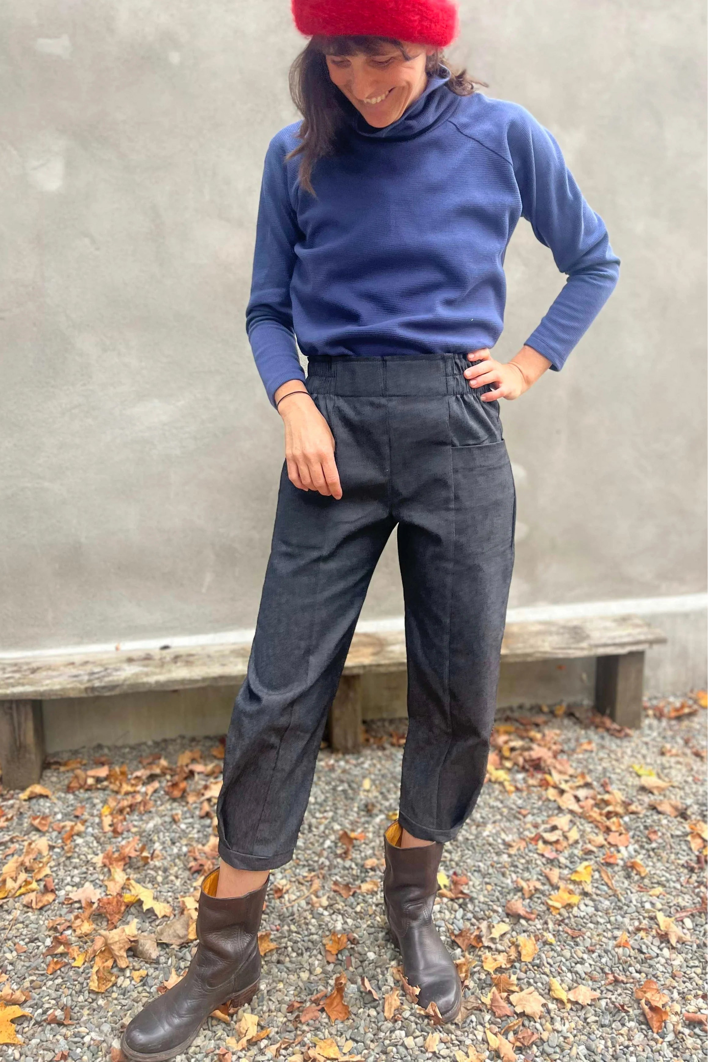 A woman wearing the Nyssa Pants by Kazak in Black Denim, barrel-legged pants with an elastic waistband and patch pockets. She is wearing them with the Oris Turtleneck in Indigo Waffle and standing outside by a grey wall. 