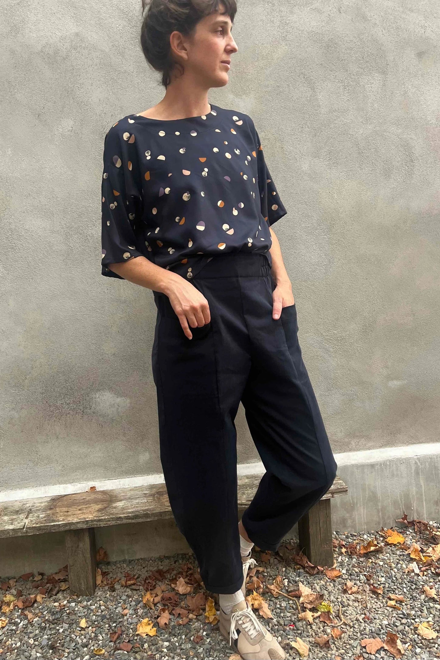 A woman wearing the Nyssa Pants by Kazak in Navy, barrel-legged pants with an elastic waistband and patch pockets. She is wearing them with the 
Abelia Top in Midnight Moons and standing outside by a grey wall. 