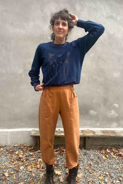 A woman wearing the Nyssa Pants by Kazak in Terracotta, barrel-legged pants with an elastic waistband and patch pockets. She is wearing them with the Marilou Crewneck in Navy and standing outside by a grey wall. 